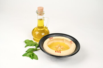Traditional Middle Eastern Hummus Dish with Olive Oil, Chickpeas, and Paprika in a Black Bowl, Accompanied by Fresh Mint Leaves and a Glass Bottle of Olive Oil on a White Background