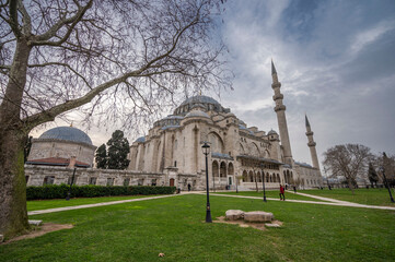 Majestic view of the S&uuml;leymaniye Mosque in Istanbul with its grand domes and towering minarets under a cloudy sky, surrounded by a peaceful garden and winter-bare trees
