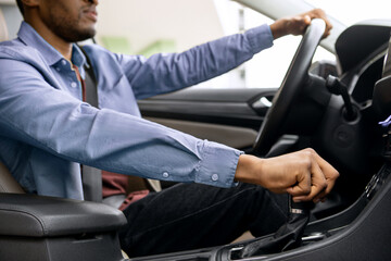 Closeup shot of black man holding steering wheel and gear shift looking at road, sitting in modern vehicle, driving his new auto