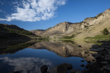 Fototapeta premium Still lake mirrors mountain, trees, sky, and clouds on a sunny day