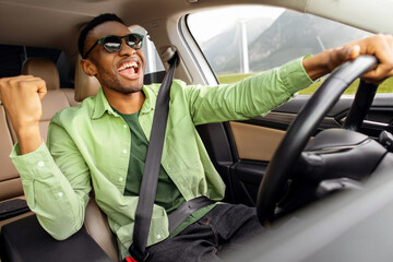 Overjoyed African American guy in sunglasses dancing in car, listening music and singing while...
