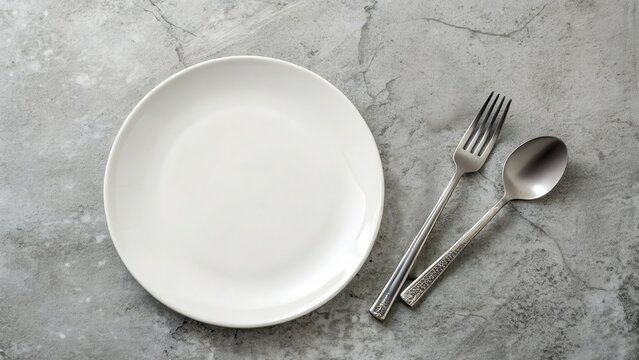 Top view of a plain white ceramic plate next to a silver fork and spoon with textured handles on a gray stone background.