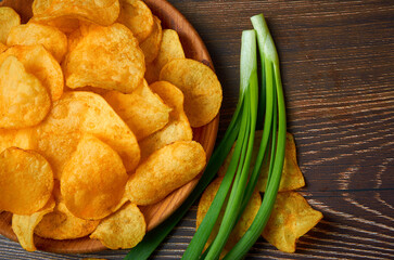 A plate of potato chips sits on a wooden table with a bunch of green onions. The chips are scattered all over the table and the onions are stacked in the background.