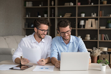 Two smiling young brothers identical twins businessmen entrepreneurs students collaborate on paperwork at home office desk make research analyze marketing stats prepare project using notebook tablet.