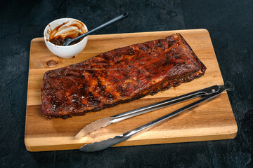St. Louis Cut Ribs auf Holzbrett, Holzteller mit Grillzange, Sch&uuml;ssel mit Barbecue Sauce und Pinsel auf schwarzem Schiefer