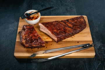 St. Louis Cut Ribs auf Holzbrett, Holzteller mit Grillzange, Sch&uuml;ssel mit Barbecue Sauce und Pinsel auf schwarzem Schiefer