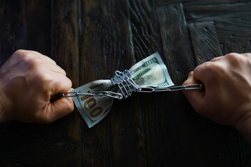 Hands Clenching Chains Around Dollar Bills on Dark Wooden Surface - Symbolic Representation of...