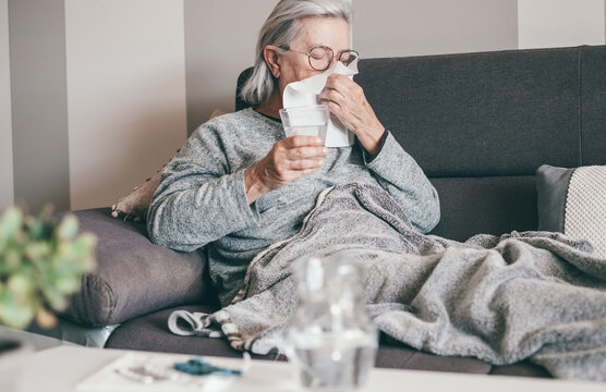 Senior caucasian woman with severe cold and fever symptoms like seasonal flu or covid blows her nose - woman on sofa at home with blanket - Powered by Adobe