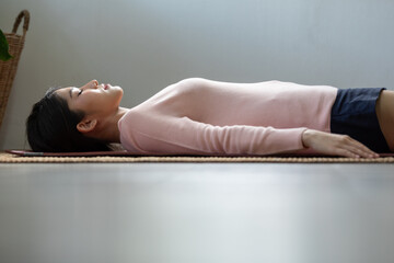 Asian woman lying on her back on yoga mat, wearing pink sweater and black shorts, in calm and...