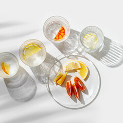 Citrus Infused Water in clear Glasses casting artistic light reflections and palm leaf shadows at sunlight. Summer drink concept, minimal pattern, flavored water with yellow red slices of fruit
