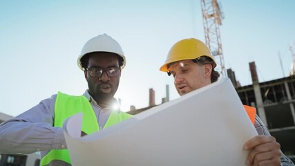 Two male engineers in helmets and vests reviewing a construction blueprint at a building site. Focused discussion with cranes and structural framework visible in the background. Outdoor teamwork.