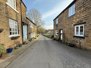 Obraz premium Charming stone houses line Church Lane, as this narrow street leads past leafy trees in the distance. The rustic scene is complemented by potted plants close to the cottages in, Esholt, Yorkshire, UK