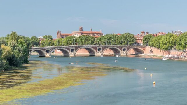Garonne River and Pont Neuf timelapse in downtown Toulouse, France. Renaissance arch bridge reflects in the water under a blue sky with Basilica of Our Lady of the Daurade. Waterfront with green trees