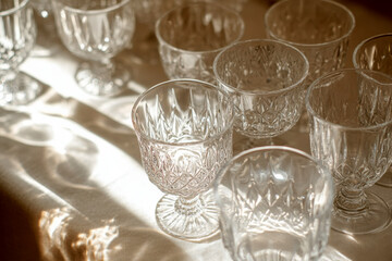 A collection of empty crystal glasses with ornate patterns sits on a table bathed in soft sunlight.