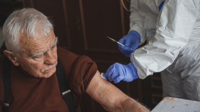 Doctor in protective suit vaccinating elderly man at home during epidemic
