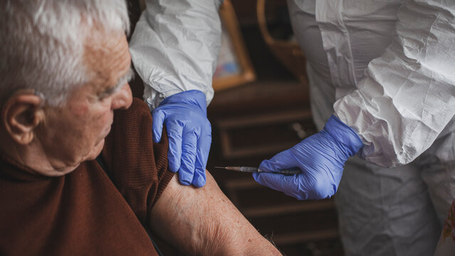 Doctor in protective suit vaccinating elderly man at home during epidemic - Powered by Adobe