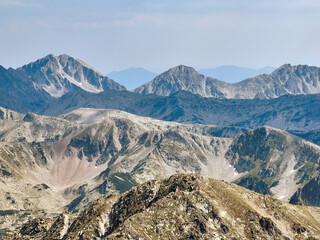 Aerial image of Pirin Mountains, Bulgaria, jagged peaks cutting through summer sky, rocky terrain and barren alpine ridges bathed in midday light, clouds drifting peacefully above. Travel