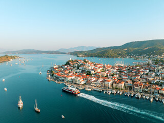 Sunset over Poros Island Greece aerial view drone capture of harbor. Sailing ferry with tourists. Mediterranean town with red rooftops, calm blue sea boats in marina, hills. Summer vacation concept
