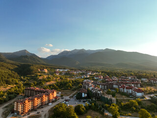 Mountain resort town with red-roofed lodges, scattered buildings, dense woodlands. Rolling hills, winding trails, serene natural surroundings enhance tranquil alpine charm. Bansko, Bulgaria. Travel