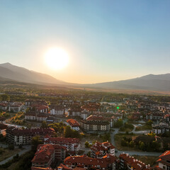 Mountain resort town with red-roofed lodges, scattered buildings, dense woodlands. Rolling hills, winding trails, serene natural surroundings enhance tranquil alpine charm. Bansko, Bulgaria. Travel