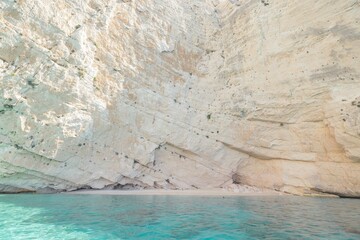 Bright summer sunshine illuminates the majestic cliffs surrounding a tranquil beach in Greece. Crystal-clear waters gently lap at the shoreline, creating a serene atmosphere