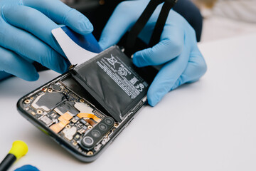 Battery replacement in a mobile phone – a technician in protective gloves disassembles a smartphone, removing the back cover to install a new battery. Repair and maintenance process of electronics.