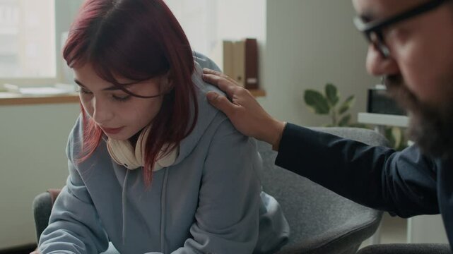 Chest up shot of upset teenage Caucasian girl with red hair and headset on neck telling about worries and emotional pain and receiving support from professional who listening and patting her shoulder