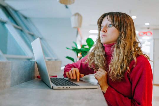 Young businesswoman with wide opened eyes looking surprised while reading shocking news on laptop, sitting in modern coworking space, experiencing astonishment and disbelief