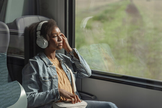 Young African American woman comfortably sitting by the train window, watching the beautiful scenery and enjoying music through her headphones. - Powered by Adobe