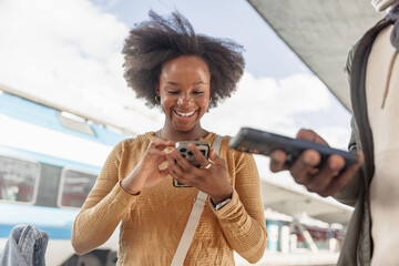 Smiling African American tourist couple with luggage using smartphone while waiting for a train on...