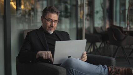 Happy middle age businessman using laptop computer in office. Older casual man working online on terrace of cafe, smiling.
