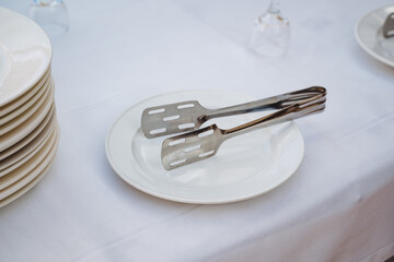 A single tong is resting on a pristine white plate that is placed on the surface of a table,...