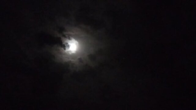 The full moon appears from behind the clouds moving across the gloomy sky. Clouds move across the moon. The full moon tries to peek out from behind the big clouds at night