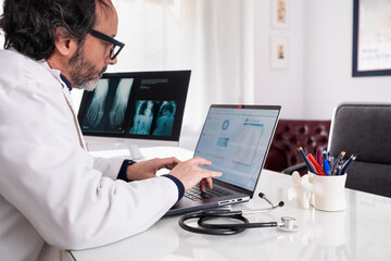 A middle-aged doctor wearing a white coat types on his laptop using generic anonymous healthcare...