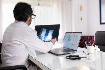 A middle-aged doctor wearing a white coat examines an X-ray. On his laptop, there is generic anonymous healthcare software. In the background, another screen shows X-rays and a corridor.