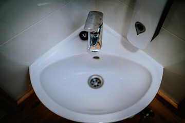 Top view of a white corner sink with a chrome faucet and wall-mounted soap dispenser in a clean tiled bathroom or washroom space..