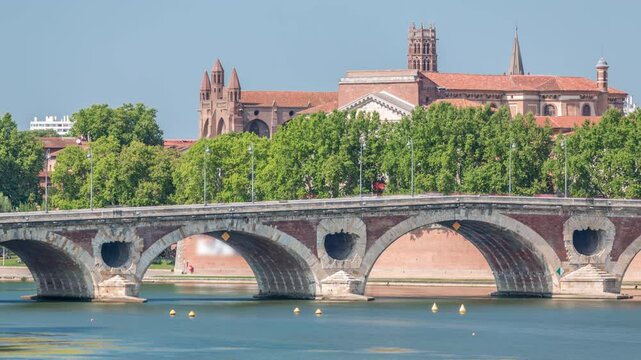 Garonne River and Pont Neuf timelapse with Basilica of Our Lady of the Daurade in downtown Toulouse, France. Renaissance arch bridge reflects in the water under a blue sky. Waterfront with green trees
