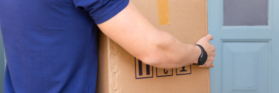 Close up cropped view of unknown male courier carrying large cardboard parcel box standing in front of addressee door. Reliable package delivery services, logistics, and electronic commerce fulfilment