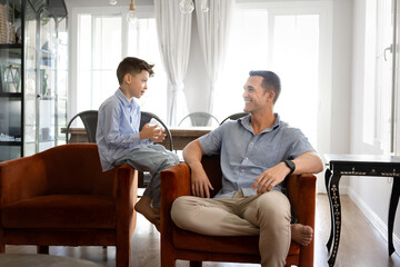 Young loving dad and little 8s son engage in meaningful conversation on leisure in living room,...