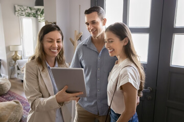 Real estate agent presenting information on tablet to smiling couple standing modern flat hallway. Woman realtor provide professional services to clients related to property buying, renting, or repair