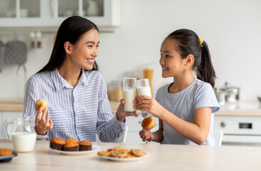 Happy asian girl and her mother drinking milk while sitting in kitchen, clinking glasses and...