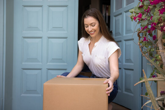 Happy woman sat down to pick up parcel box left by courier on porch, smiling, enjoy convenience of modern doorstep ordered package delivery services. Customer satisfaction, trust and positive feedback
