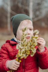 Young child holding a bouquet of fluffy willow branches in early spring, wearing a red jacket and green hat, standing outdoors in a natural rural setting with a blurred background.