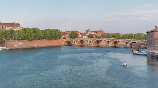 Garonne River and Pont Neuf timelapse with Port de la Daurade in downtown Toulouse, France. This Renaissance arch bridge reflects in the water under a blue sky with clouds. Waterfront with green trees