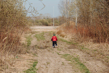 A child in a red jacket and green hat rides a blue balance bike on a paved path next to a field. The child is facing away from the camera, and the field in the background is brown and barren.
