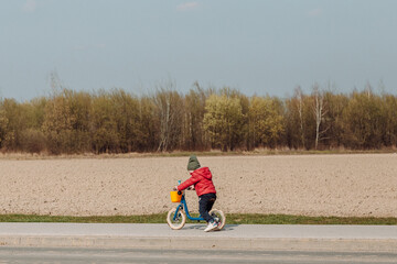A child in a red jacket and green hat rides a blue balance bike on a paved path next to a field. The child is facing away from the camera, and the field in the background is brown and barren.