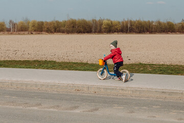 A child in a red jacket and green hat rides a blue balance bike on a paved path next to a field. The child is facing away from the camera, and the field in the background is brown and barren.