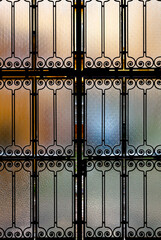 The metal gate with frosted glass closes off the entrance to the courtyard of an old city building. Through the bright sunbeams, we can see blurred spots of different shades behind the gate
