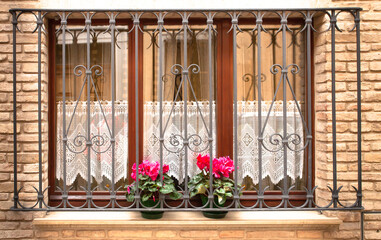 brick wall with three-section window featuring metal grillwork and two red flowers in jars.These flowers on sill along with the white curtain in window create sense of comfort and look truly amazing