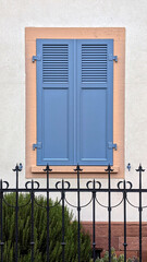 Closed window by blue wooden shutters on a light-colored wall and ornate iron fence detail in a...